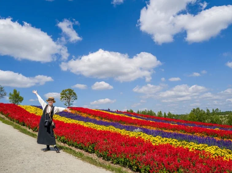 麦わら帽子をかぶった女性が、赤、黄、紫のカラフルなストライプ状の花畑で両腕を広げて笑顔を見せている