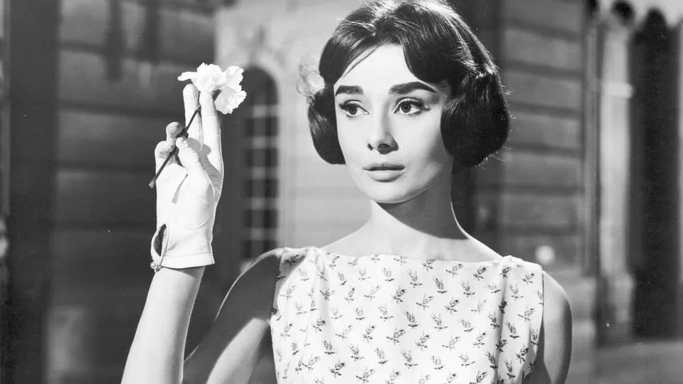 白いグローブをはめた手で花を持つオードリー・ヘプバーンのモノクロポートレート
