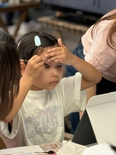 幼い女の子が真剣な表情で眉にメイクを施している様子です。白いTシャツを着ており、そのTシャツには「CEOSI」という文字がプリントされています。