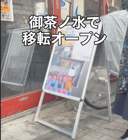 御茶ノ水での店舗移転オープンを告知するA型看板が写っています。看板にはアイドルのようなグループが写ったポスターと「安安」の文字があり、左側の壁には営業時間や料金に関する情報が書かれた貼り紙も見えます。