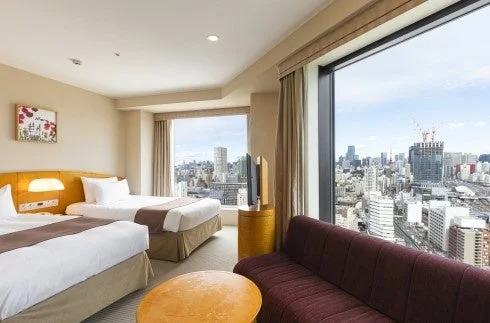 高層階にあるツインベッドのホテル客室で、大きな窓からは広々とした都市の景色が一望できます。室内にはソファとテレビも備え付けられています。