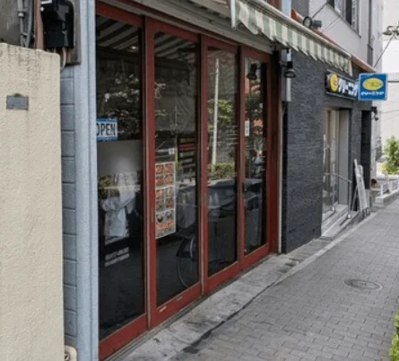 狭い通りに面した2軒の店舗の外観です。手前の店は赤い枠のガラス扉で「OPEN」の看板があり、隣はクリーニング店の黄色い看板が見えます。