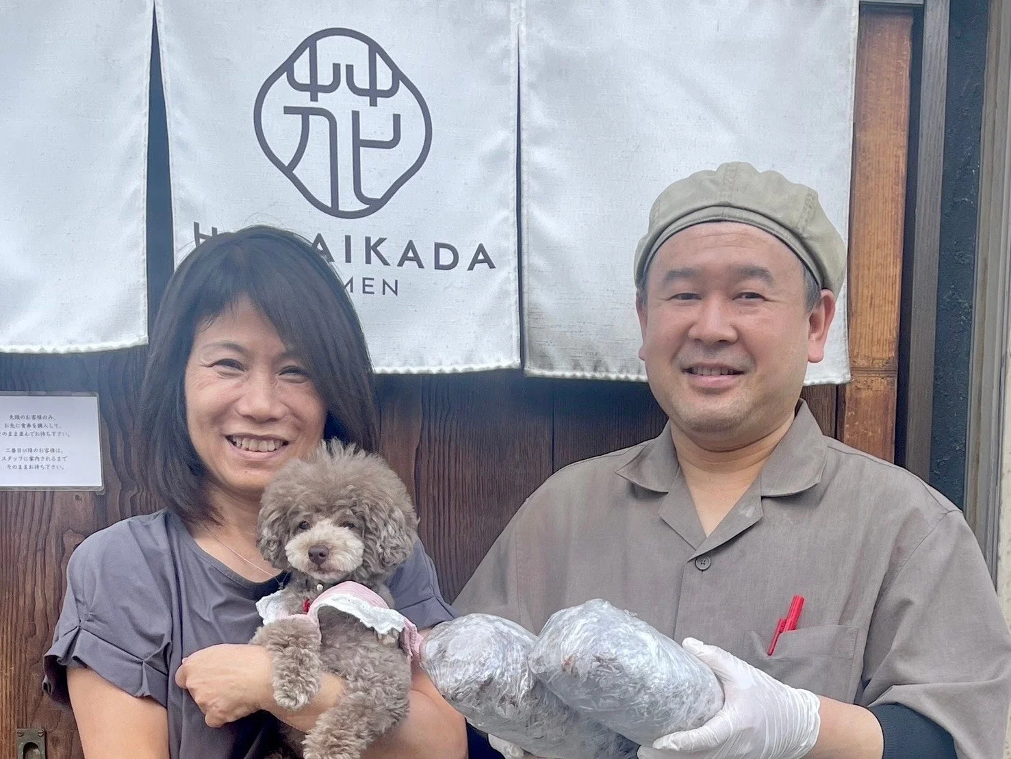 店先ののれんの前で笑顔の男女と可愛らしい犬