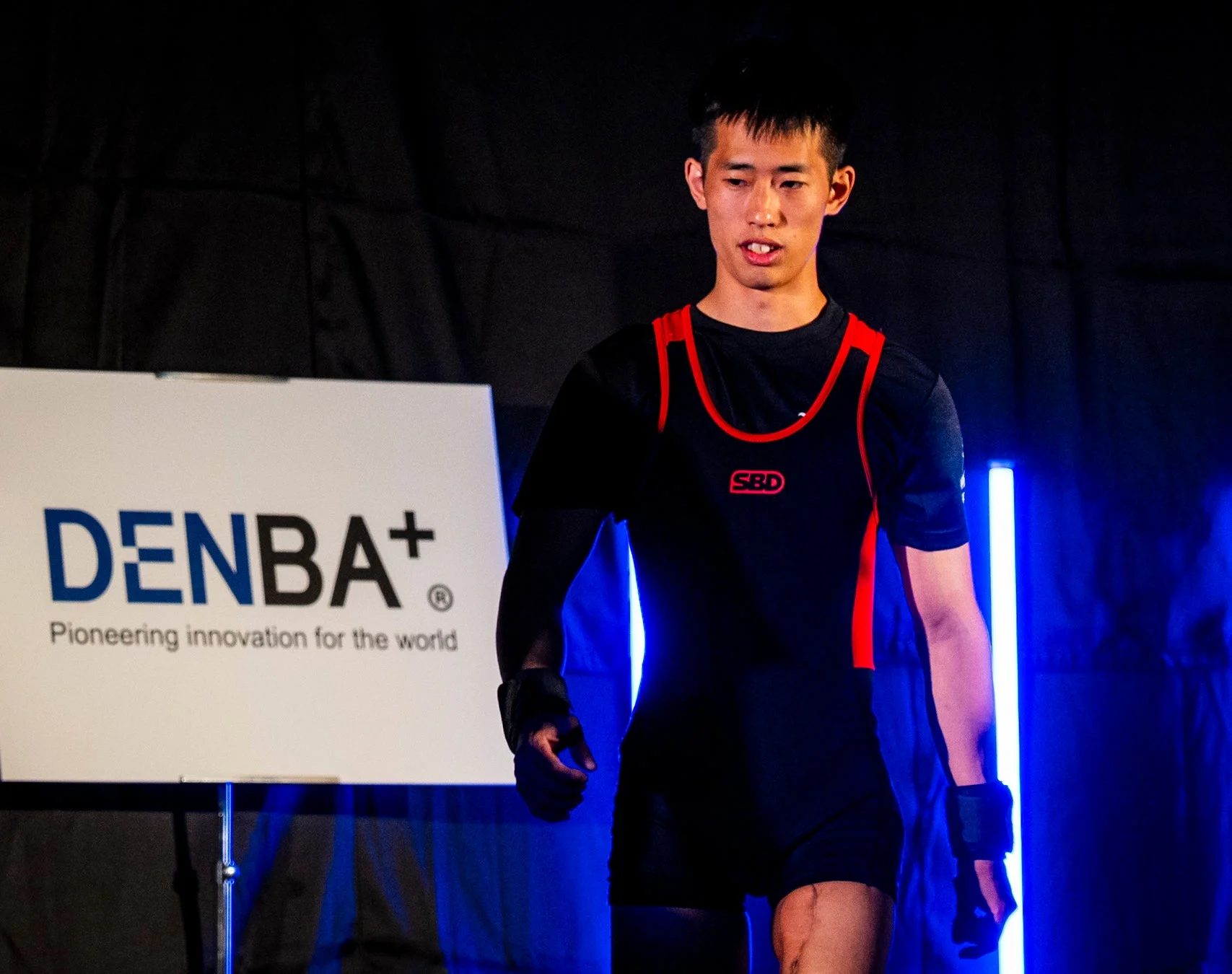 黒いスポーツウェアを着たアジア人男性アスリートが、SBDのロゴが見えるユニフォーム姿でステージを歩いている。背景には「DENBA+」の看板と青い照明がある。