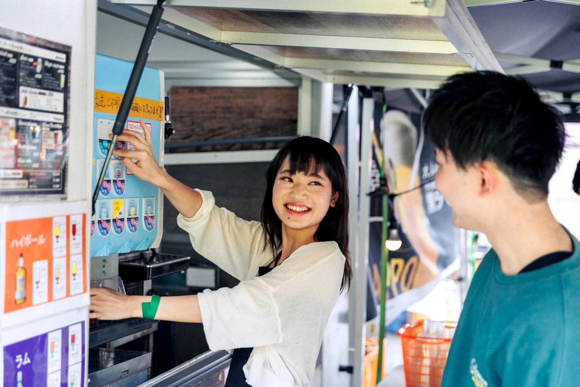 女性が笑顔でドリンクディスペンサーを操作し、男性と楽しそうに交流している様子。屋外のドリンクスタンドで、賑やかな雰囲気が伝わってきます。