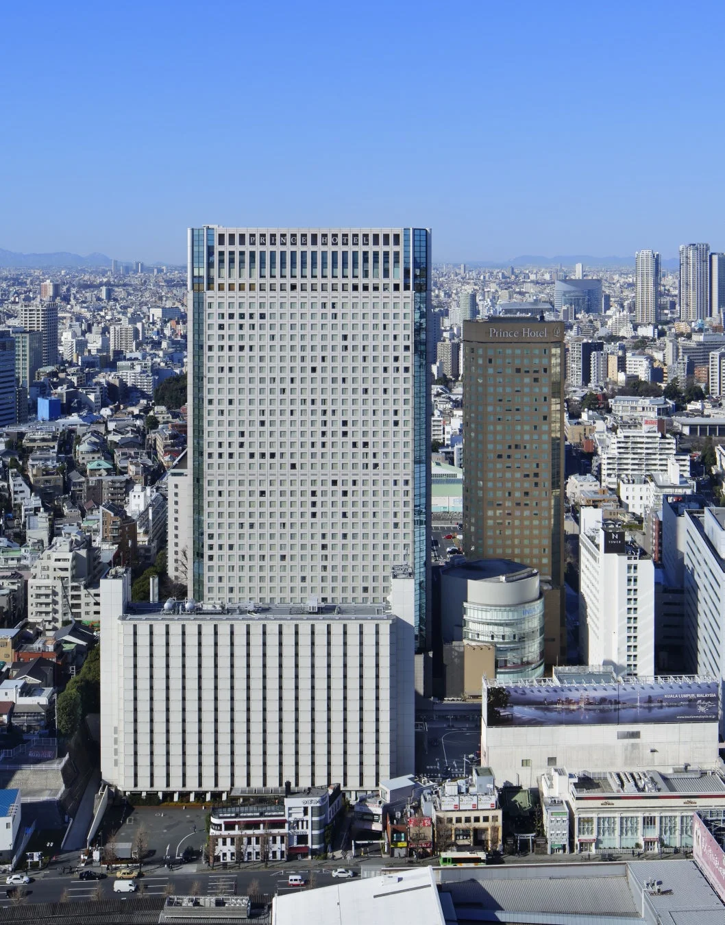 晴れた青空の下、高層ビルが密集する都市の俯瞰風景です。中央に「PRINCE HOTEL」の文字が見える大型ビルがあり、奥にもプリンスホテル系の建物が見えます。遠景にはクアラルンプール、マレーシアの広告看板があります。