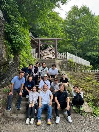 緑豊かな自然に囲まれた石段で、約15人ほどのグループが集合写真を撮っています。背景には鳥居のある日本の伝統的な建物が見え、和やかな雰囲気で旅行や観光を楽しんでいる様子が伺えます。