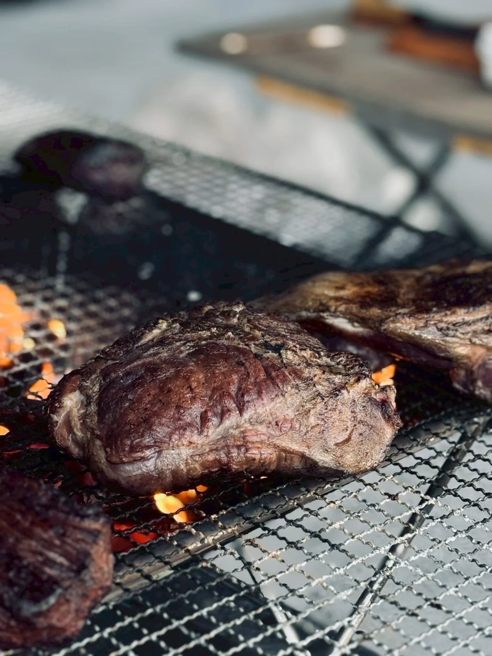 炭火で焼かれている大きな肉の塊が写っています。網の上でじっくりと調理されており、炎が肉の下から見え、食欲をそそる様子です。アウトドアでのバーベキューやキャンプ料理を思わせる一枚です。