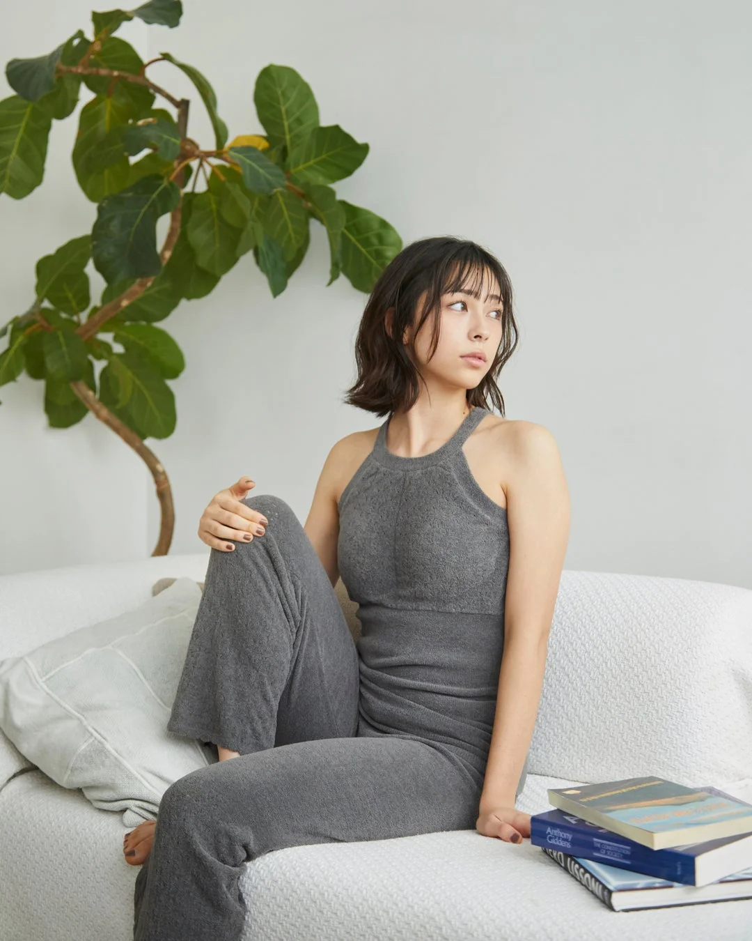 グレーのカジュアルな部屋着を着た女性が白いソファに座り、リラックスした様子で遠くを見つめています