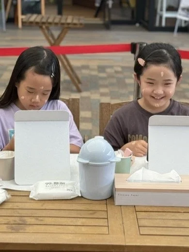 2人の女の子が屋外のテーブルでメイクアップ体験をしている様子。顔にクリームを塗ったり、鏡を見たりして楽しんでおり、特に右の女の子は笑顔を見せている。テーブルには除菌シートやティッシュが置かれている。