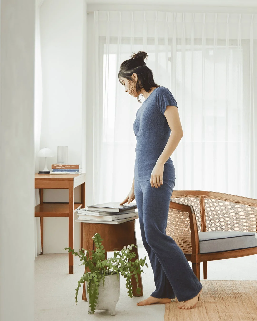 明るい部屋で、青い部屋着を着た女性が、本が積まれたスツールに手を置いて立っている様子