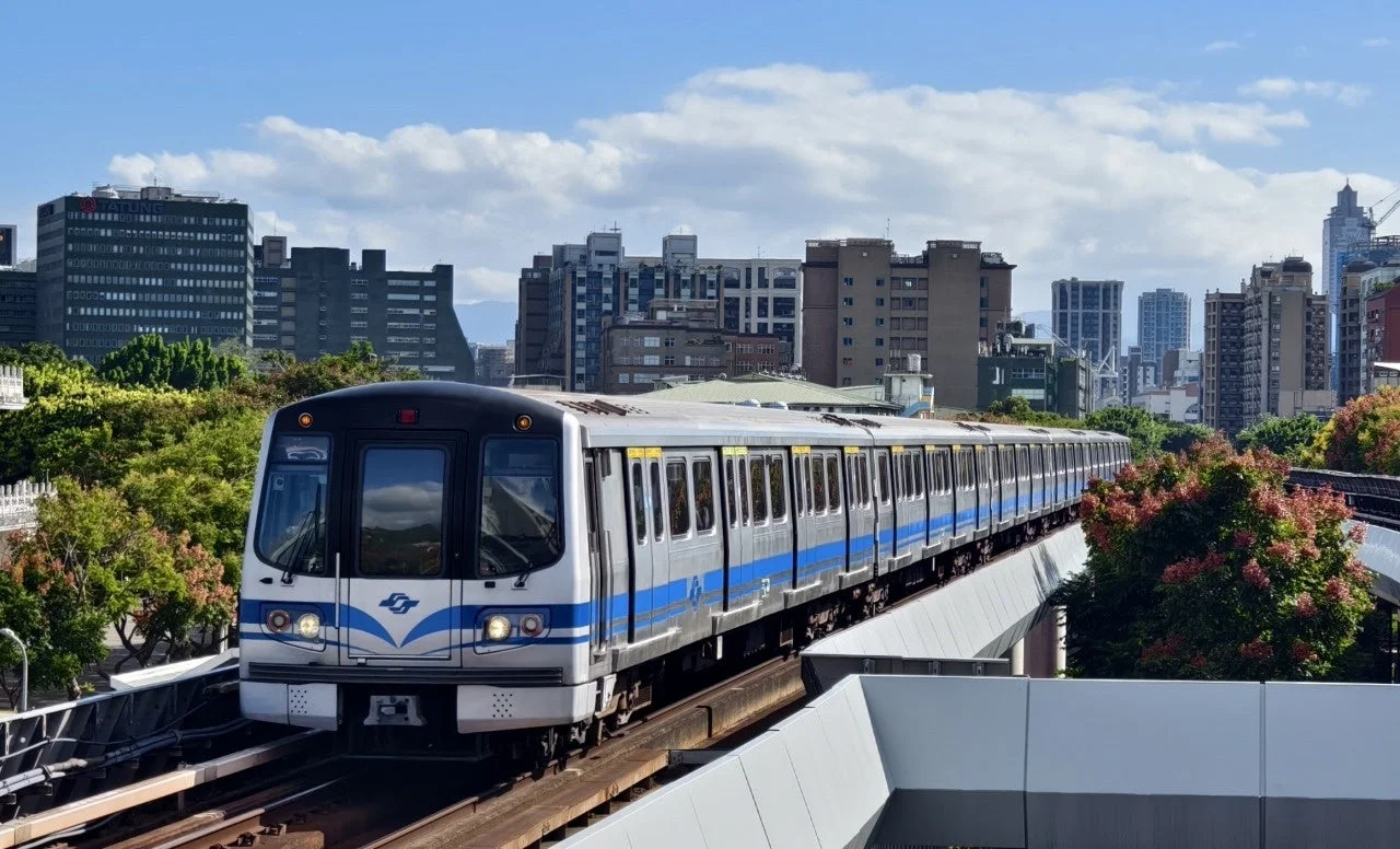 晴れた日の台北の都市風景。高架線を走る台北捷運の電車が中央に写っており、背景には高層ビル群と青い空、手前には緑豊かな木々が広がっています。
