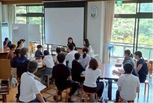 明るい室内で、大人と子供が参加する会議または学習会の様子です。二人の女性が前に立ち、プロジェクタースクリーンを背景にプレゼンテーションを行っているようです。窓からは緑が見えます。