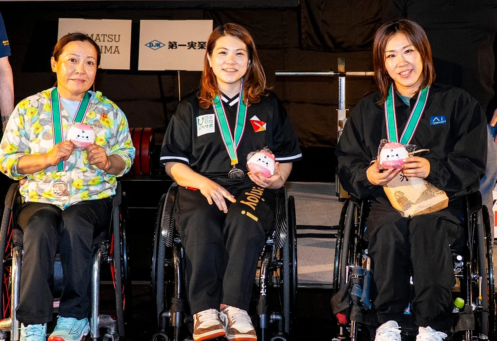 車椅子に座った3人の女性が笑顔でメダルと可愛らしいマスコットを手にしている表彰式の様子です。パラリンピックのようなスポーツイベントで、彼女たちの達成感が伝わってきます。