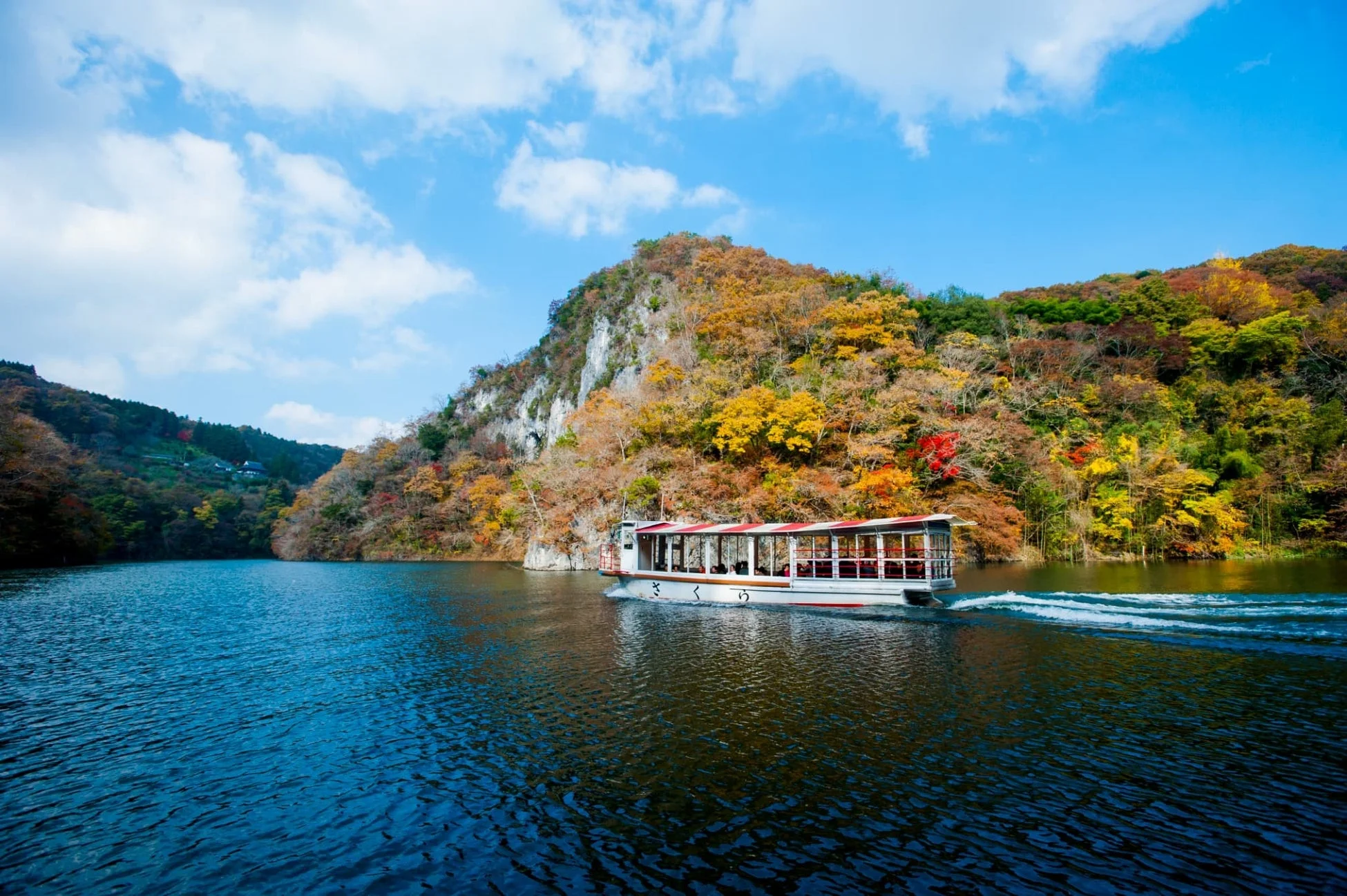 紅葉に彩られた山々を背景に、青い湖面を遊覧船が進む美しい秋の景色。