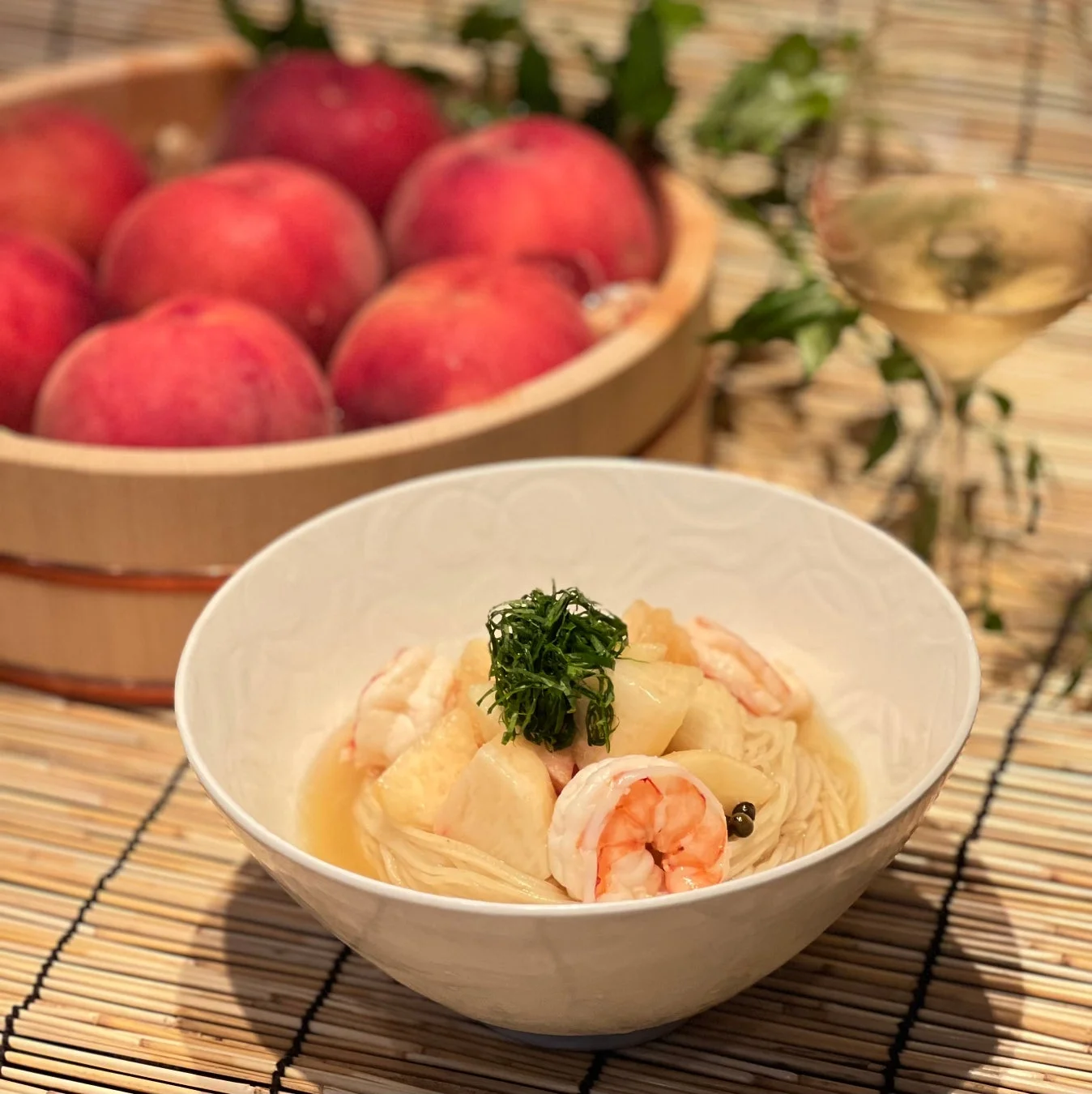 手前に海老と白い具材が乗った素麺の器があり、背景には木桶いっぱいの新鮮な桃とワイングラスが置かれています