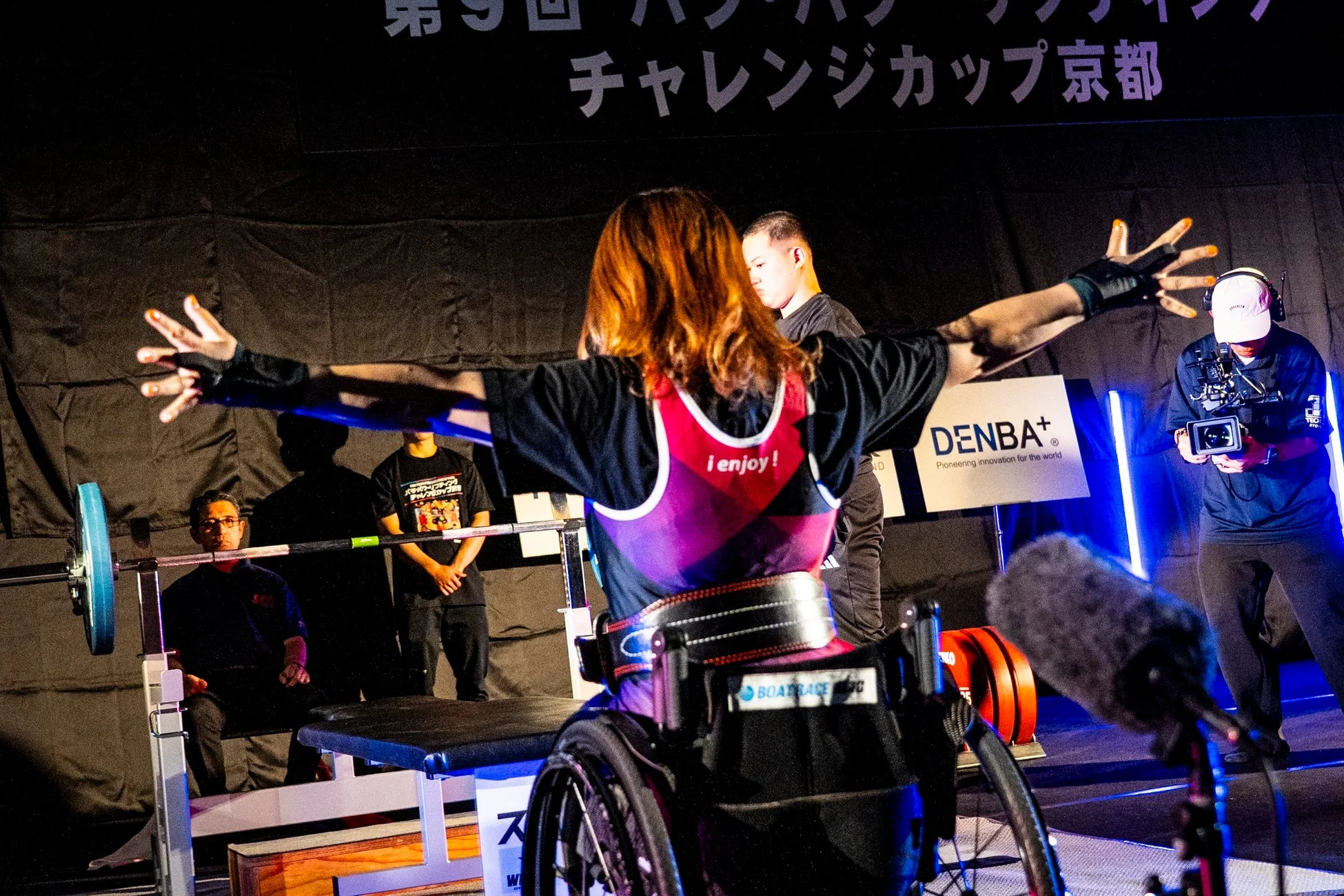 「第9回 パラ・パワーリフティング チャレンジカップ 京都」で、車いすの選手がベンチプレスに挑む瞬間を捉えた写真です。背中には「i enjoy!」の文字があり、腕を広げて集中している様子が伺えます。大会の真剣な雰囲気が伝わります。