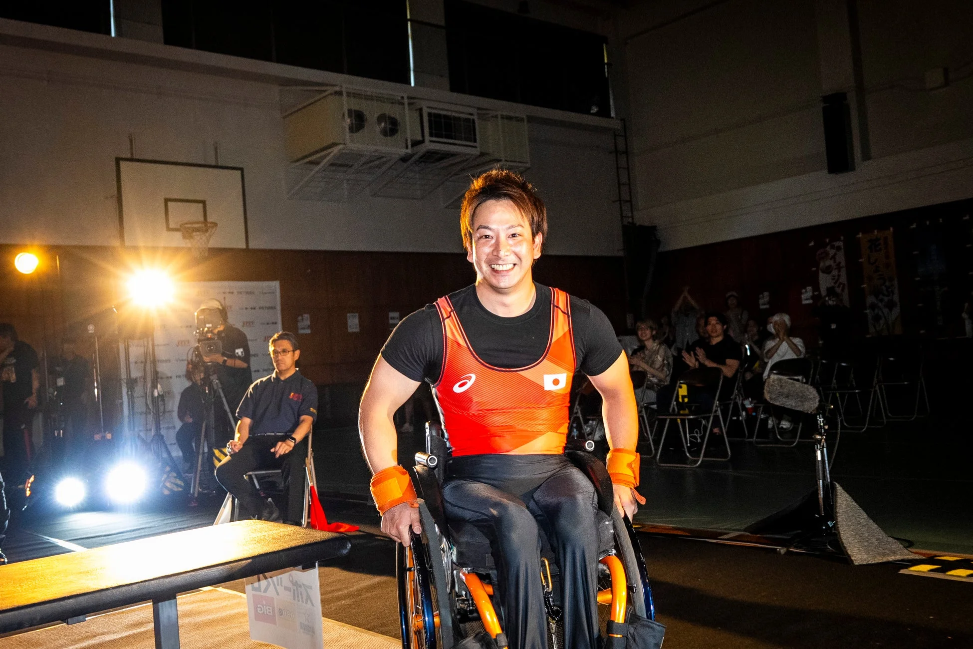 日本の国旗が付いたオレンジ色のユニフォームを着た車いすアスリートの男性が、体育館で明るい笑顔を見せている。