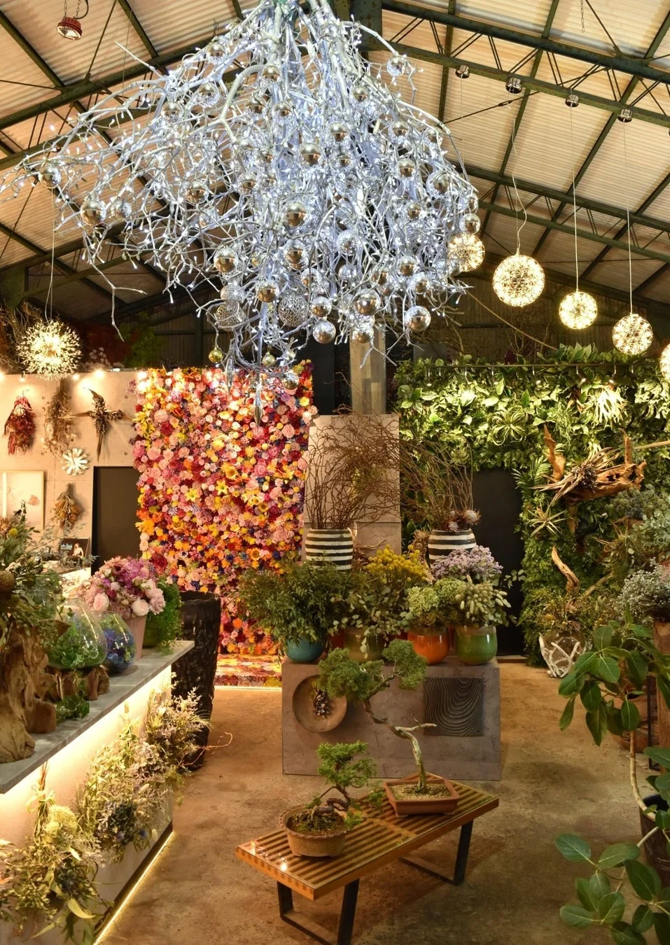華やかな装飾が施された広々とした花屋または植物園の内部。天井には枝とオーナメントでできた大きなシャンデリアが吊るされ、壁には色とりどりの花々が飾られている。様々な観葉植物や盆栽が並び、自然とアートが融合した美しい空間です。
