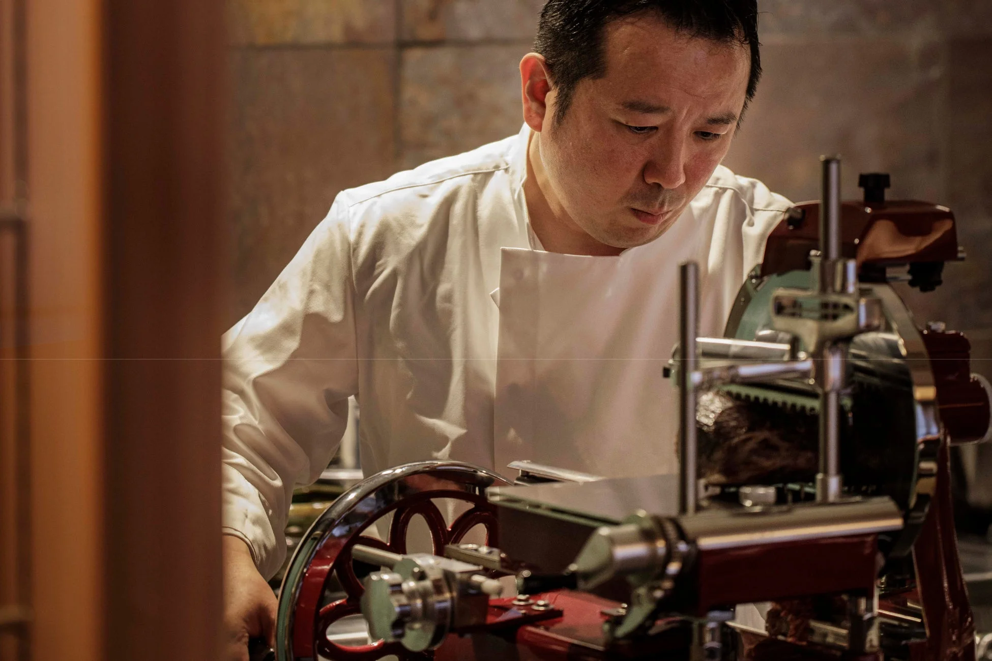 白いコックコートを着た料理人が、プロ仕様の肉スライサーを使って真剣な表情で肉をスライスしています