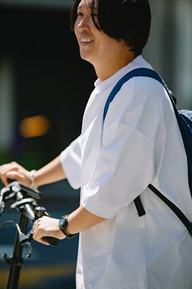 白いTシャツを着てリュックを背負った男性が、自転車のハンドルを握りながら笑顔を見せている。
