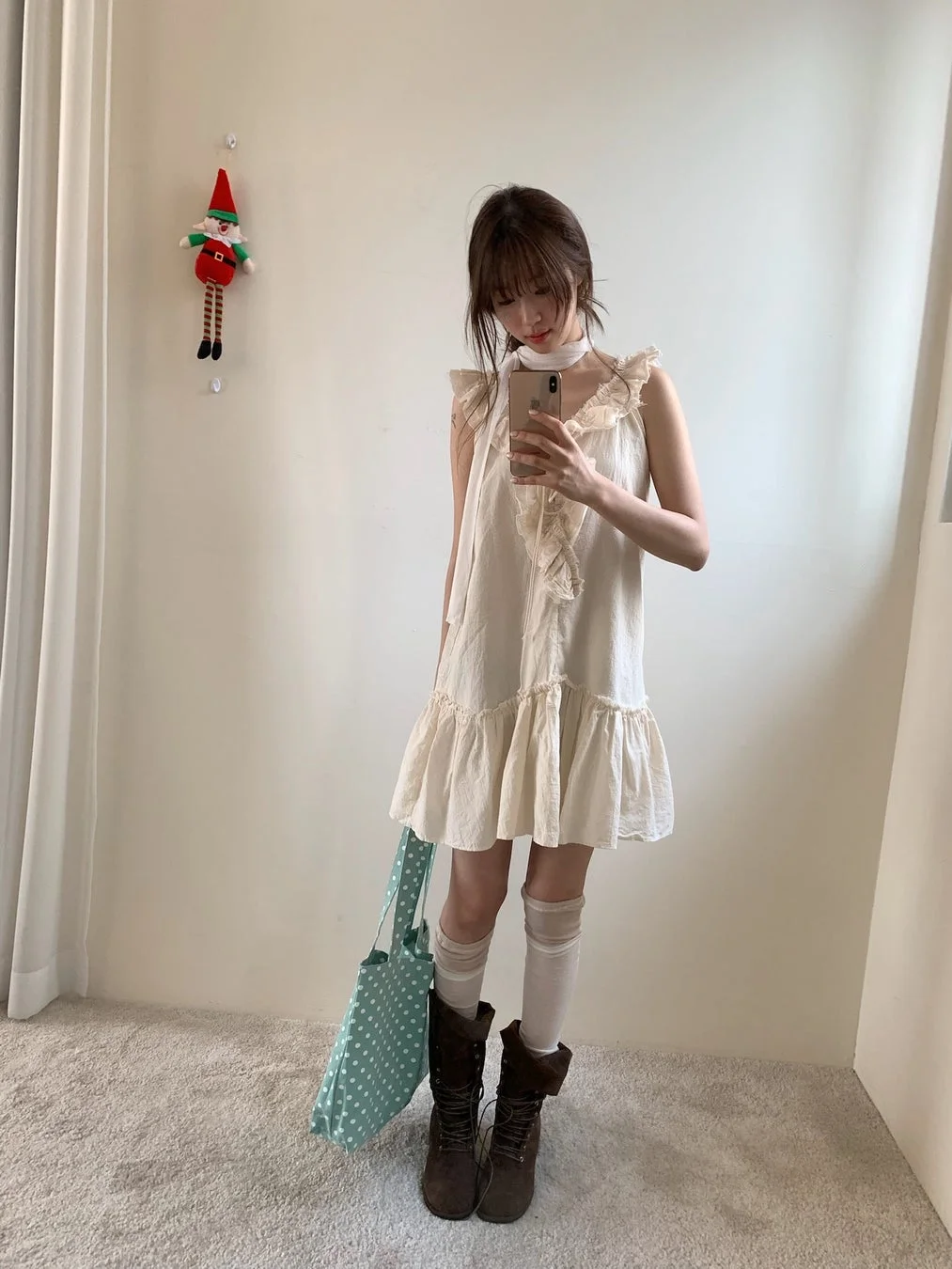 鏡の前で自撮りをする若い女性の全身像です。フリル付きのクリーム色のワンピースに白いニーハイソックスと茶色のブーツを合わせ、水色のドット柄トートバッグを持っています。