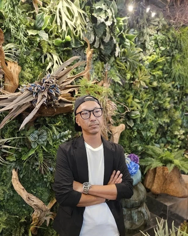 緑豊かな植物の壁を背景に、眼鏡をかけ、黒いブレザーと白いTシャツを着た男性が腕を組んで立っています。