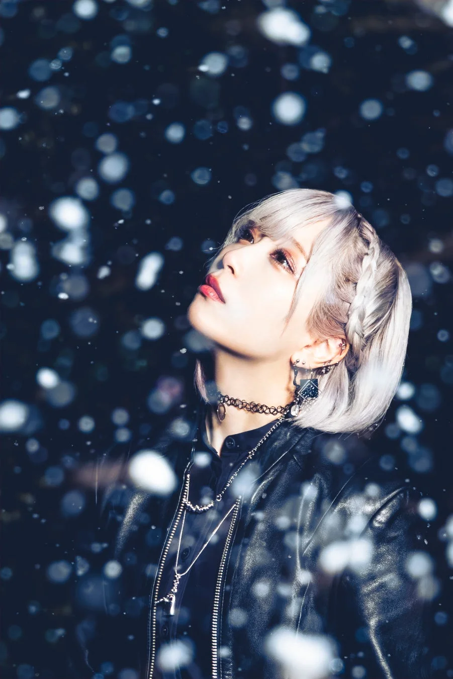 ショートのアッシュグレーの髪を編み込んだ女性が、レザージャケットを着用し、雪のような光の粒が舞う暗い背景を見上げているクールなポートレートです。