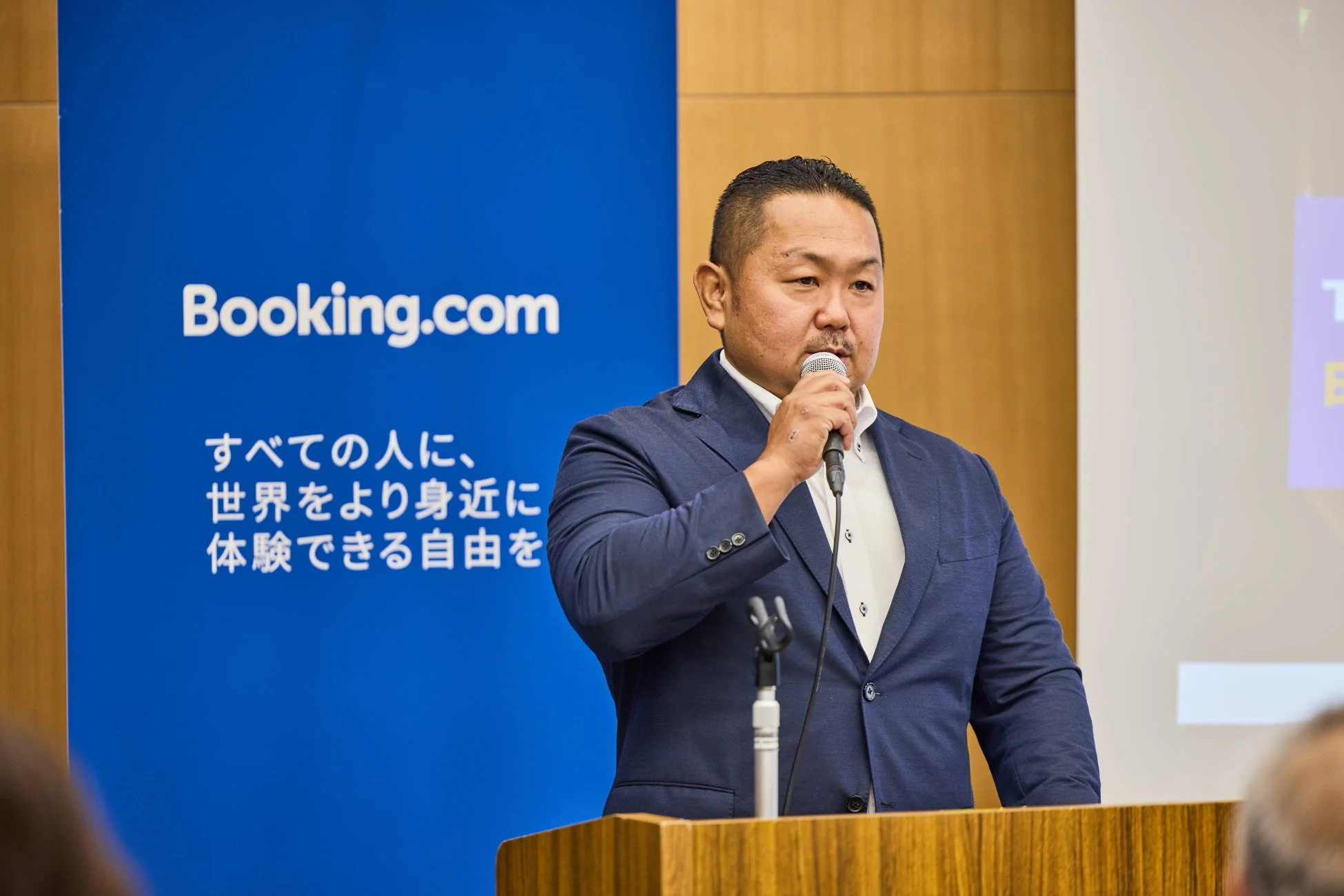 スーツ姿の男性がマイクを持って講演しており、背景には「Booking.com」のロゴとスローガンが書かれた青いバナーが見えます。ビジネスイベントでの発表の様子を捉えた写真です。