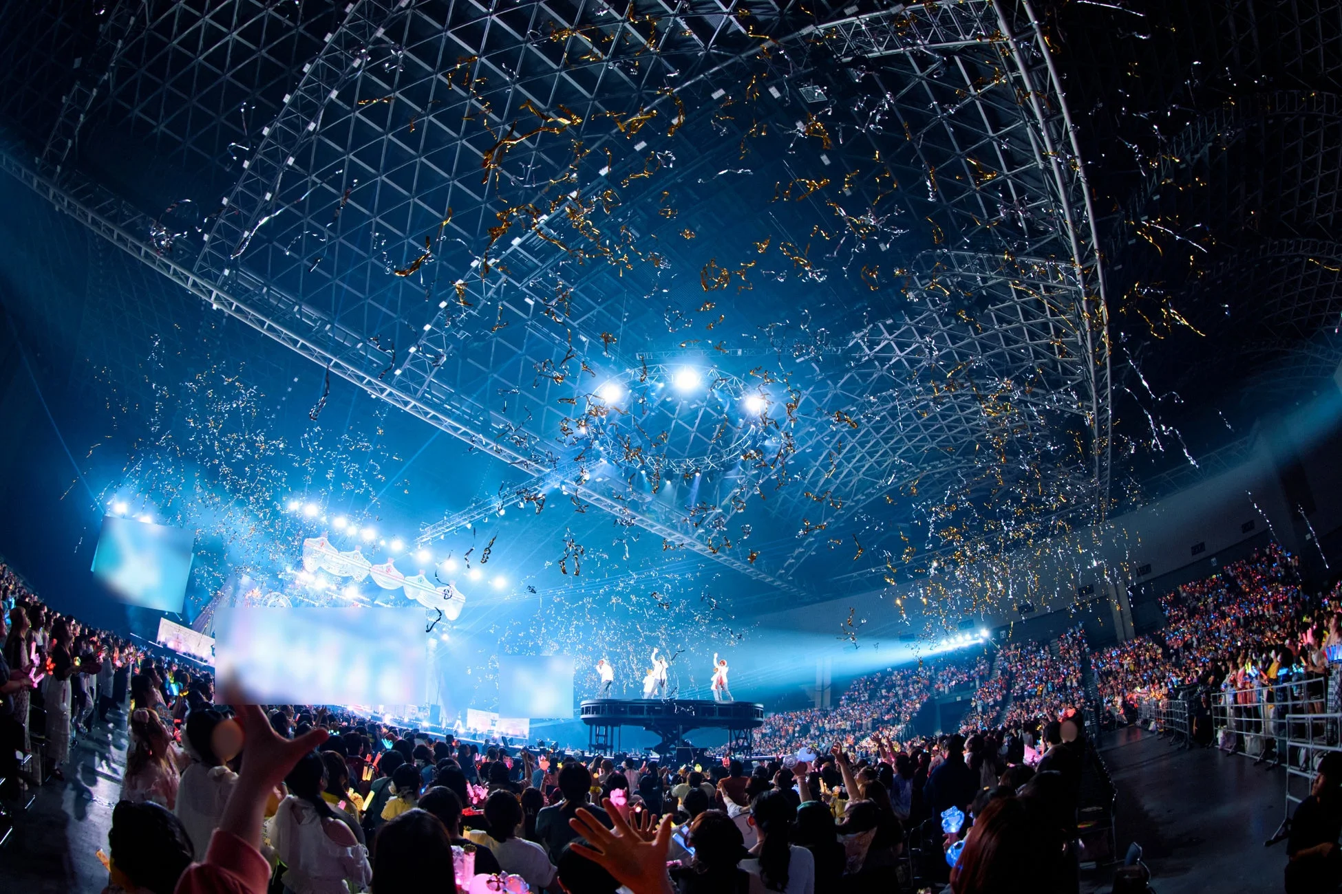 大勢の観客で埋め尽くされたコンサート会場で、ステージ上のパフォーマーが熱演しています。天井からは金色の紙吹雪が舞い、観客はペンライトを振って盛り上がっており、壮大なライブイベントの様子がうかがえます。