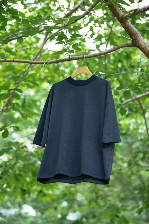 木々の緑を背景に、濃紺のオーバーサイズTシャツが木の枝に吊るされたハンガーにかかっています。