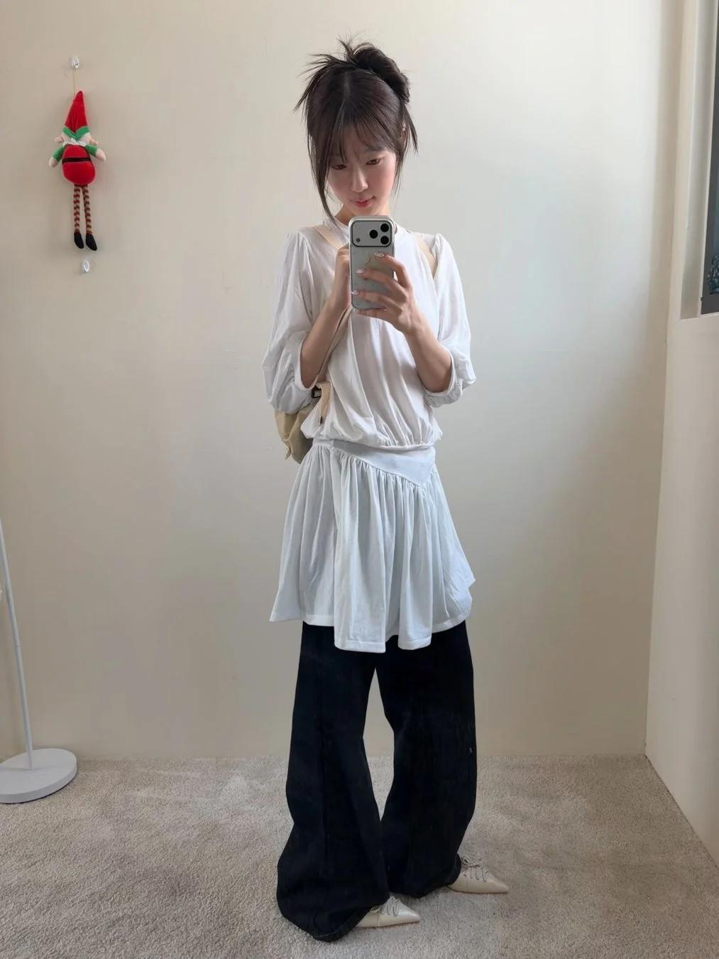 部屋で白いトップスと黒いワイドパンツを着用した若い女性が、スマートフォンで全身ミラーセルフィーを撮っています。