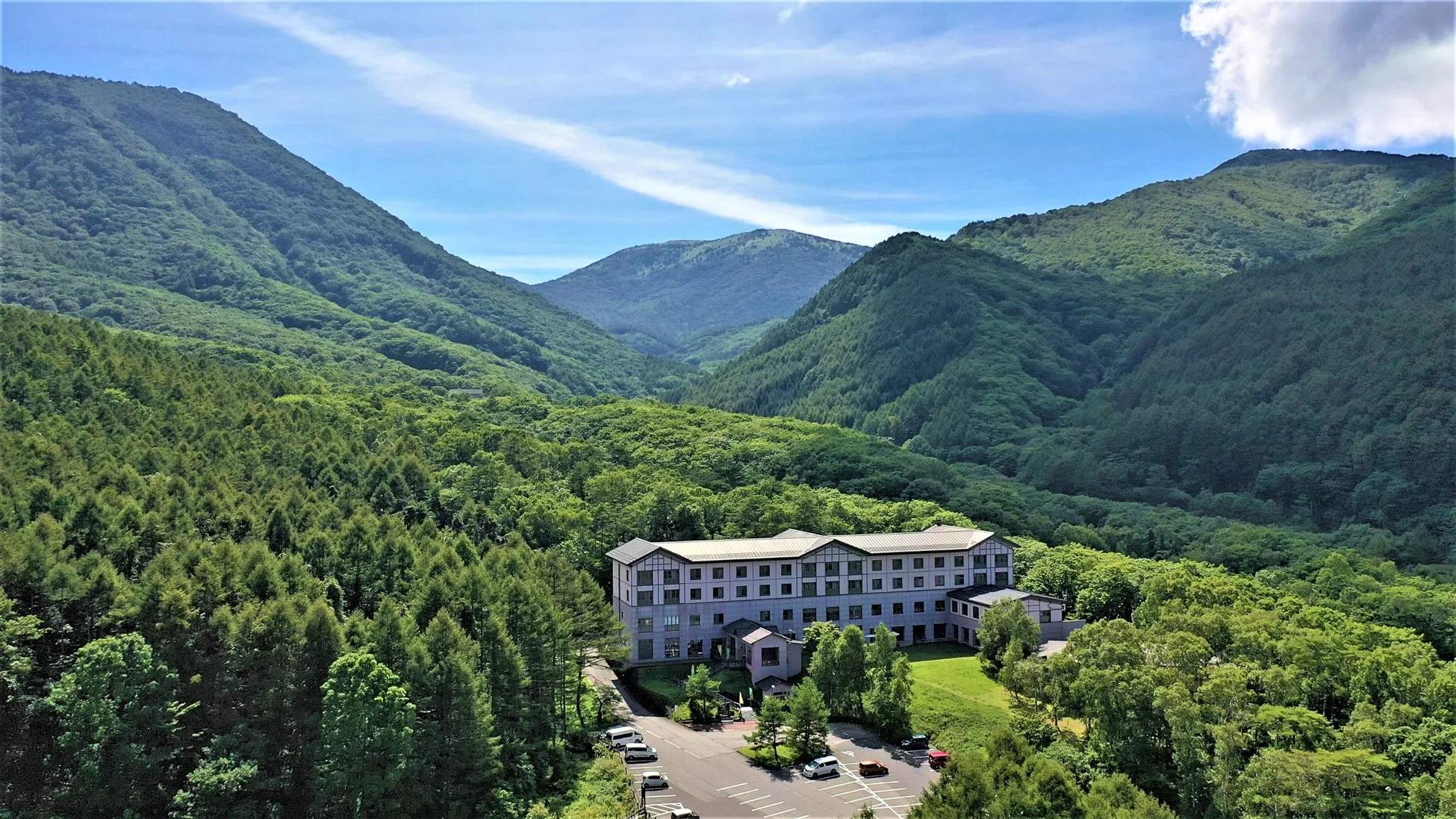緑豊かな山々に囲まれた谷間に建つ大きなホテルまたはリゾート施設の全景。晴れた青空の下、広大な自然の中に溶け込むように佇み、駐車場も見える静かで癒される風景です。