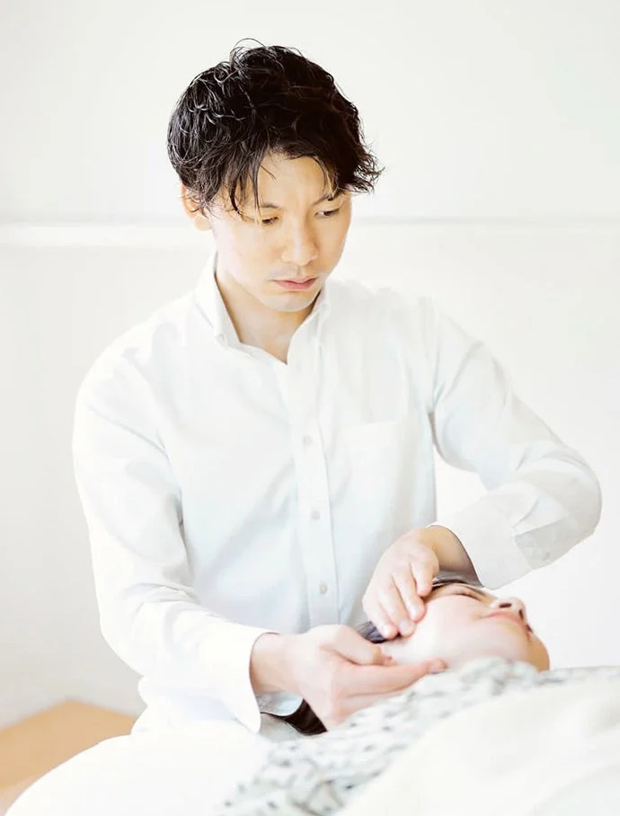 白いシャツを着た男性が、横たわる女性の顔に施術を行っています