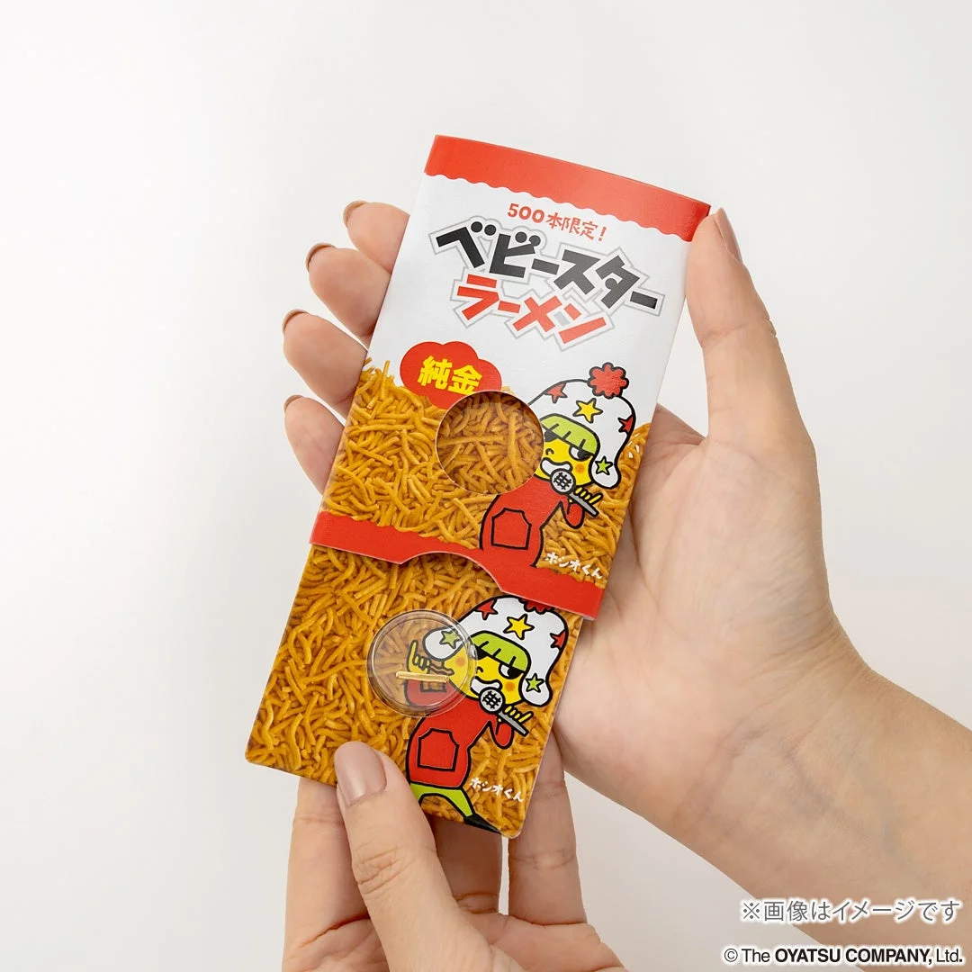 手が「ベビースターラーメン」の限定パッケージを持っている画像です。パッケージは500本限定の「純金」バージョンで、キャラクターのホシオくんが描かれています。純金製のコインのようなものが封入されているのが特徴です。