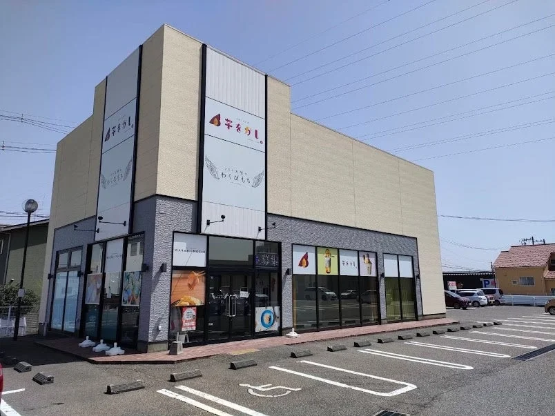 晴れた日に撮影された、和菓子店「芋をかし」の外観です。建物には「芋をかし」と「わらびもち」の大きな看板が掲げられており、店舗前には広い駐車場があります。