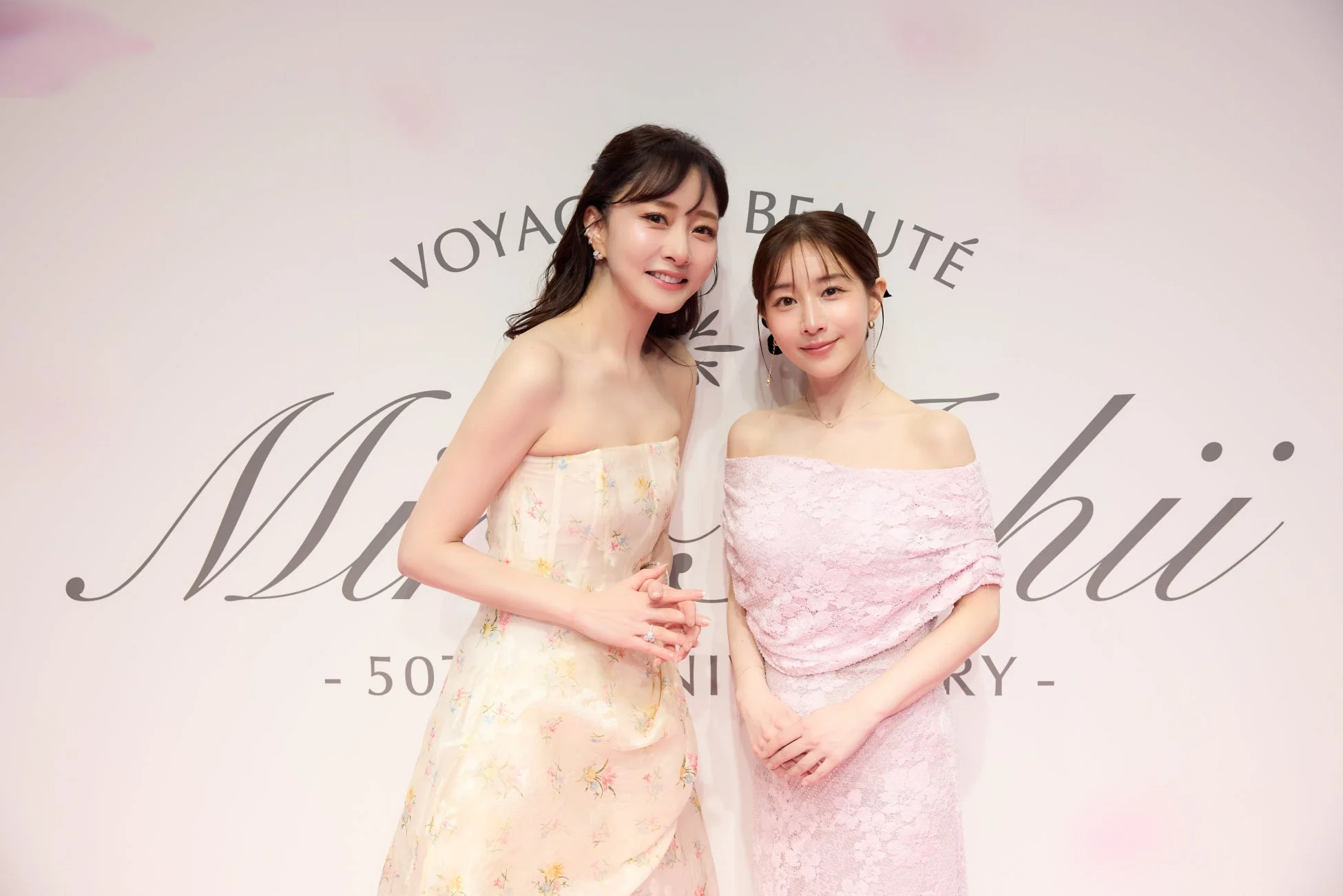 華やかなドレスをまとった二人の女性が、背景にブランドロゴらしき文字を背に笑顔で並んでいます。