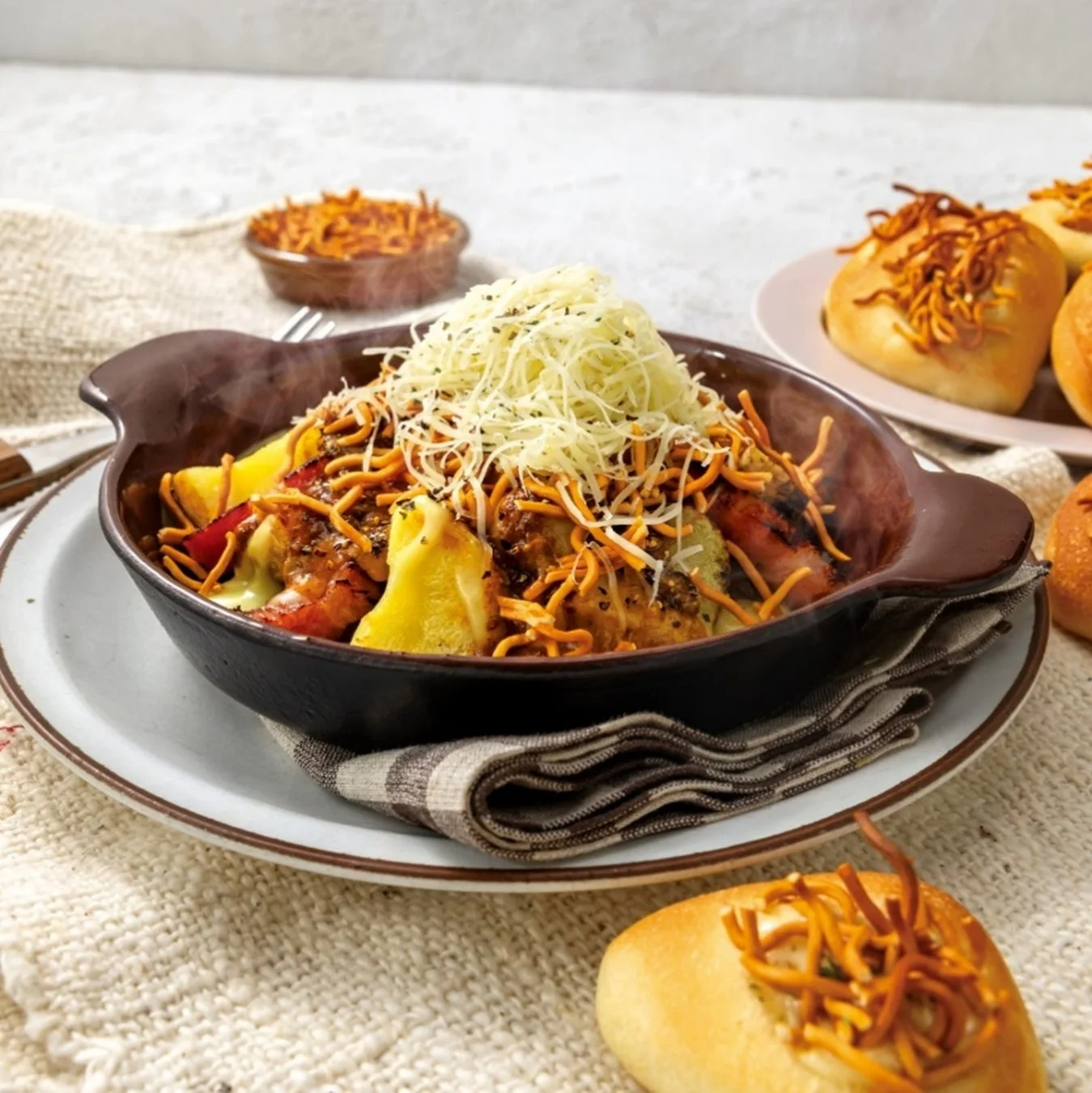 白いテーブルクロスに置かれた温かい料理とパン。中央の器には、とろけるチーズと揚げ麺がたっぷり乗ったポテトや肉の料理があり、湯気が立ち上る。背景には同じ揚げ麺がトッピングされたパンも並び、食欲をそそる一皿。