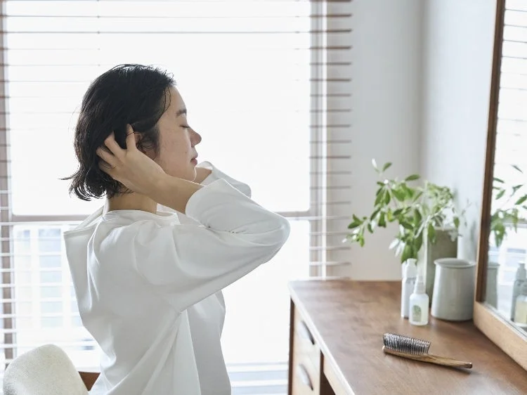 白いシャツを着た女性が、窓からの光が差し込む部屋で髪を整えている横顔の画像です。