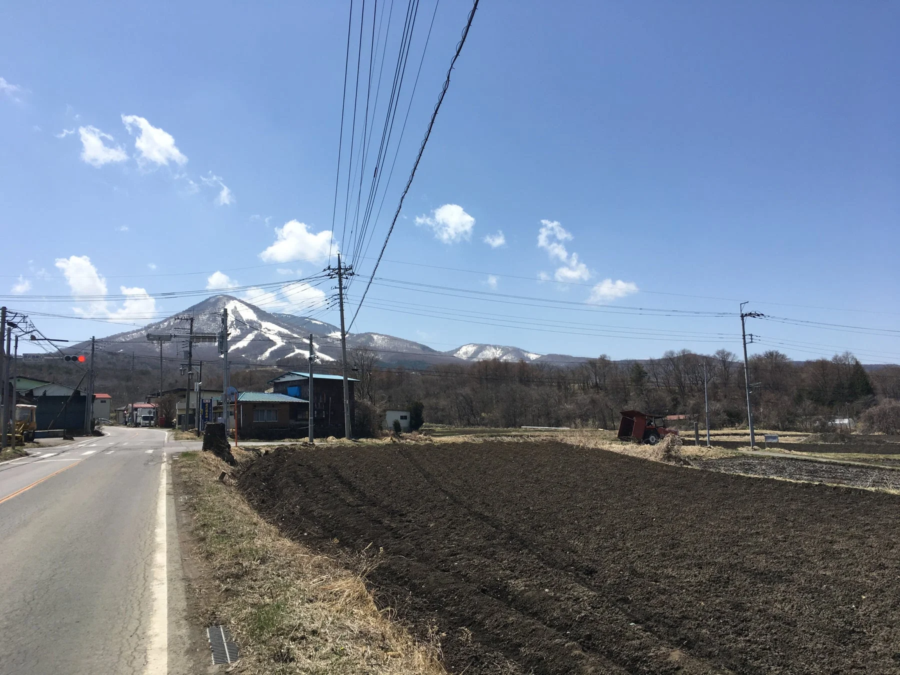 雪が残る山を背景に、耕された畑、道路、家屋が広がる日本の田舎の風景。青空には白い雲が浮かび、電線や電柱が空を横切っています。のどかな春の農村の様子を捉えた一枚です。