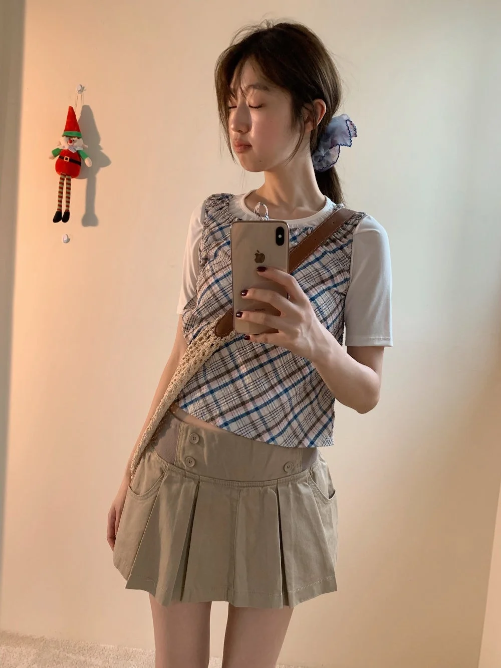 若い女性が鏡越しにスマートフォンでセルフィーを撮っています。白Tシャツにチェック柄のベスト、ベージュのプリーツミニスカートを合わせたカジュアルな服装で、肩には編み込みバッグをかけています。