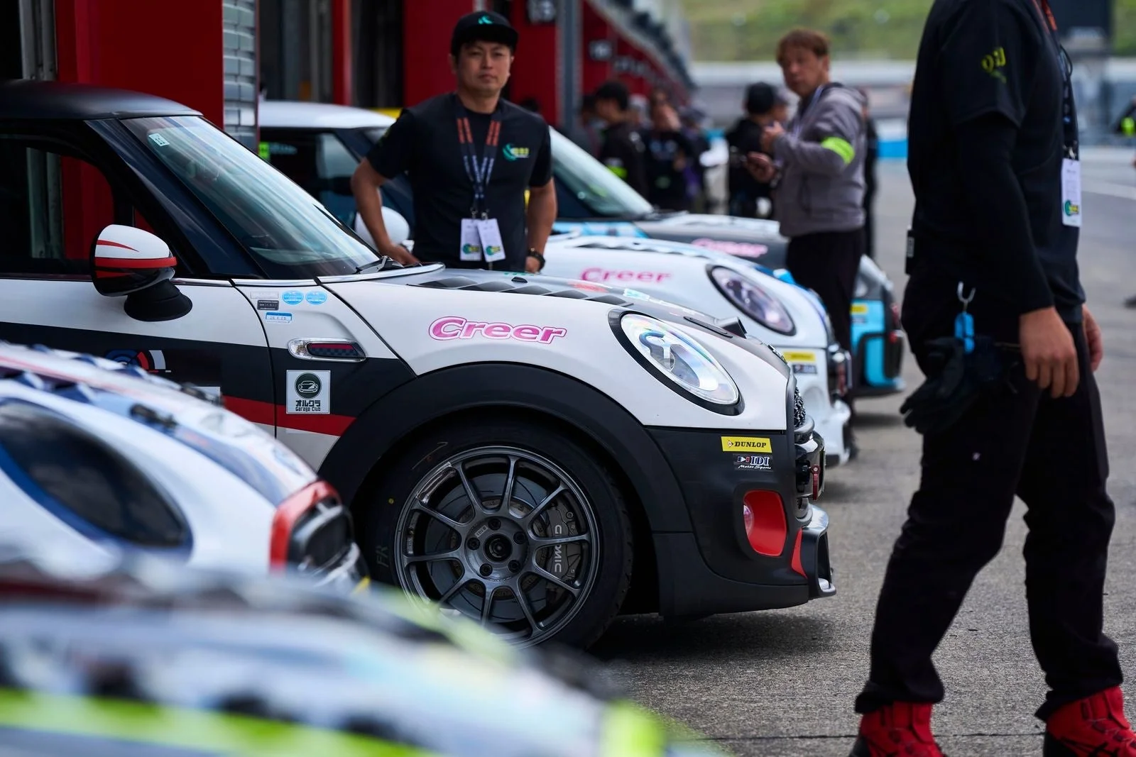 サーキットのピットレーンに並んだ複数のレーシングカーと、その周りに立つチーム関係者たちが写っています