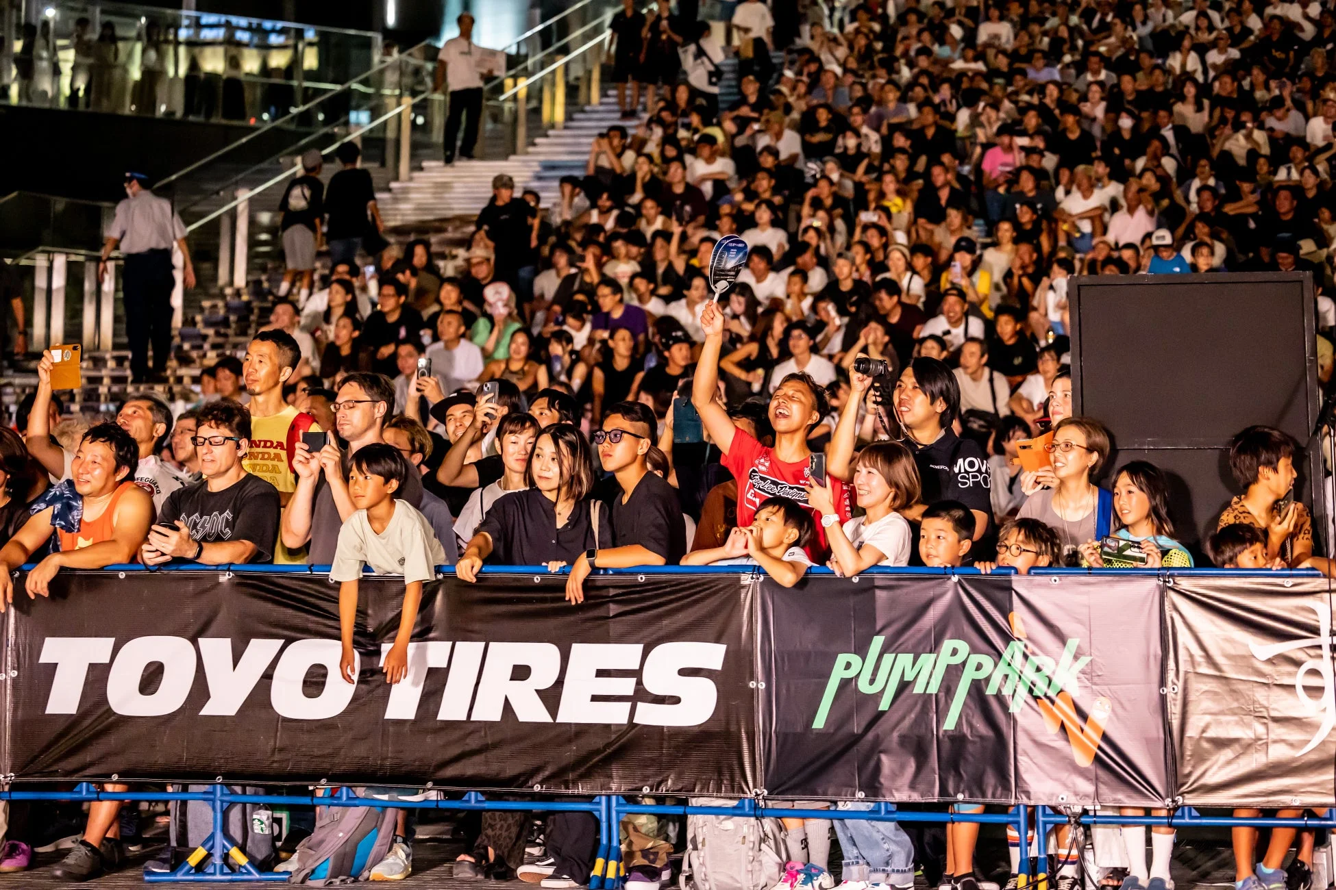 夜の屋外イベントで、多くの観客がステージを囲み、スマートフォンで撮影したり声援を送ったりしている様子