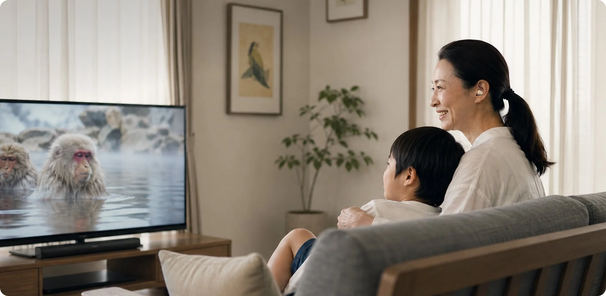 リビングのソファで、笑顔の女性と子供が一緒にテレビを見ている様子。テレビには温泉に入るサルが映っており、穏やかな家族のひとときを過ごしている。女性はイヤホンを着用している。