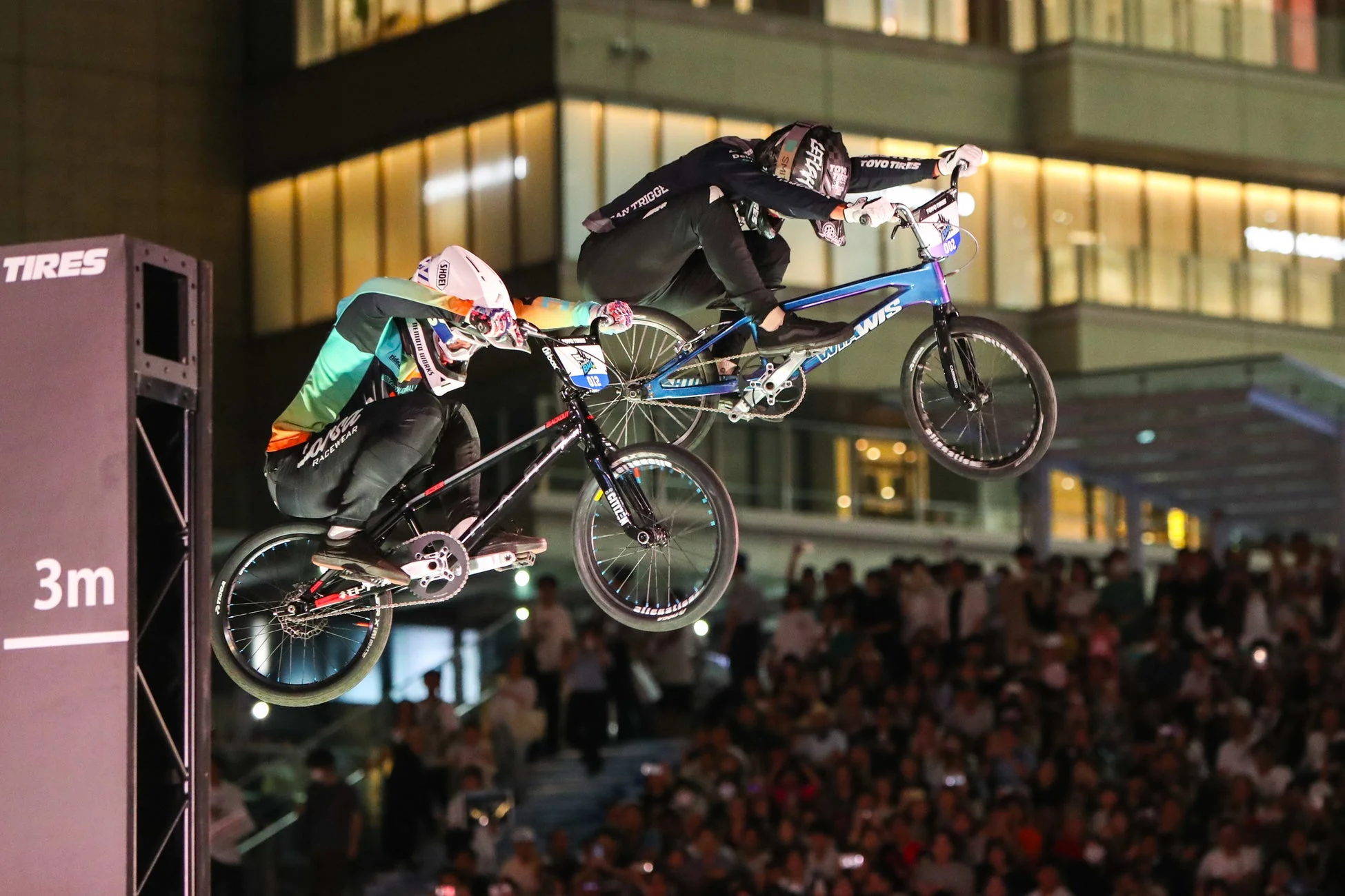 夜間のイベントで、2人のBMXライダーが空中でダイナミックなジャンプを披露