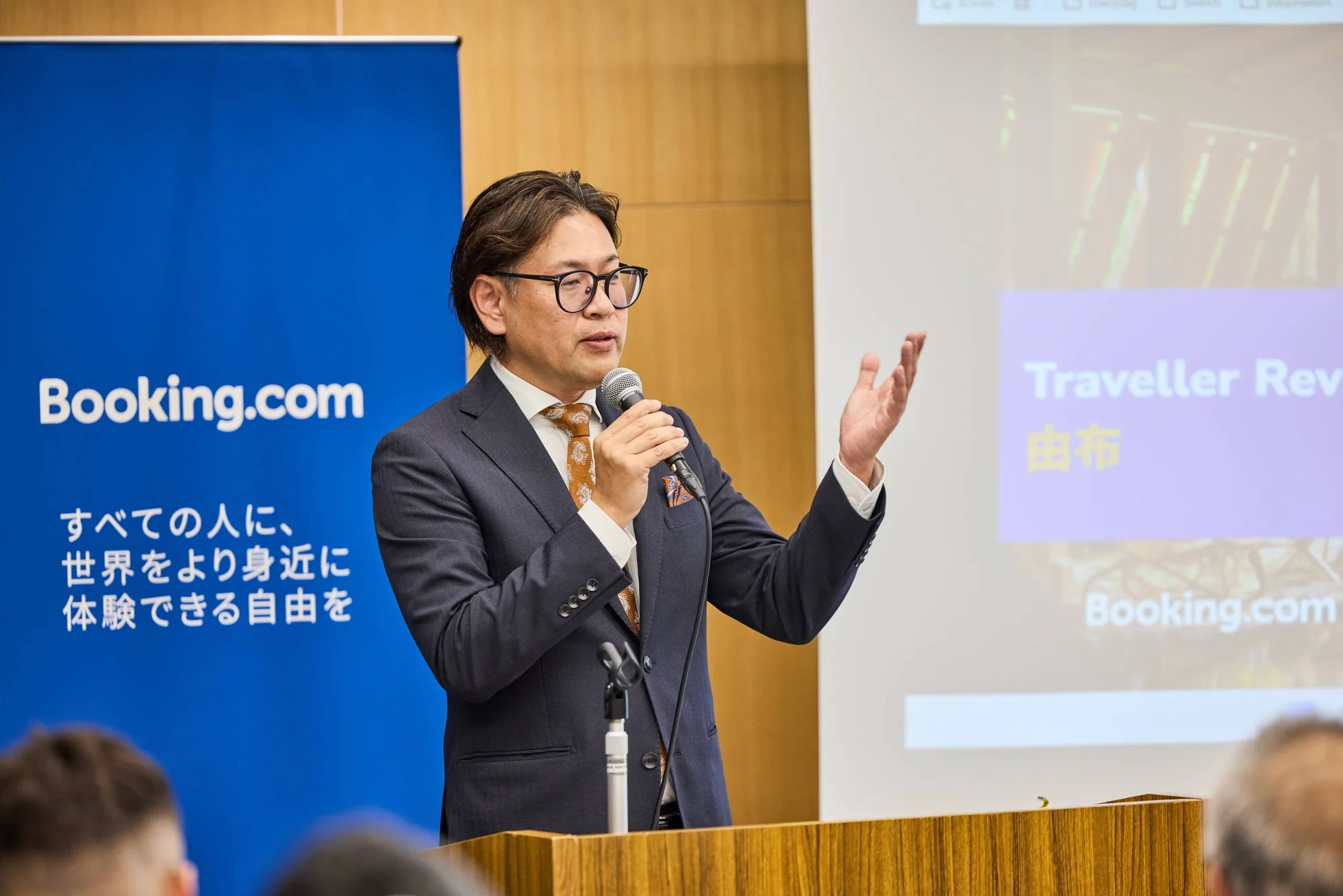 スーツを着た男性がマイクを手に講演を行っている様子。背景には「Booking.com」のロゴと「すべての人に、世界をより身近に体験できる自由を」という日本語のスローガンが書かれた青いバナーがある。スクリーンには「Traveller Rev」と「由布」の文字が表示されており、ビジネスイベントやカンファレンスの場面と見られる。