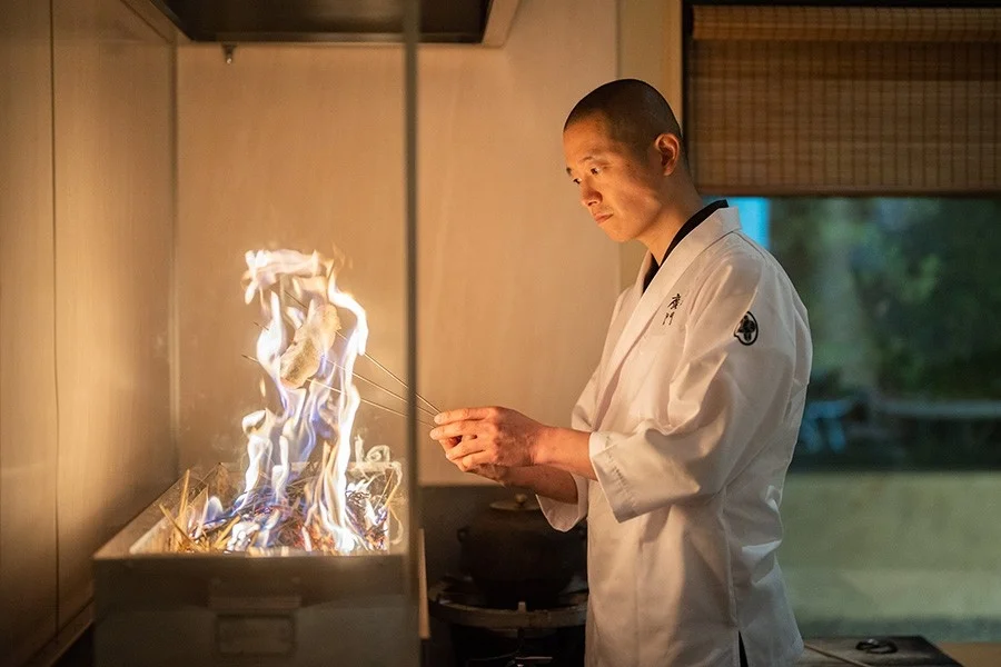 白い調理着を着た料理人が、炎が上がる炉端で串に刺した食材を丁寧に焼いています