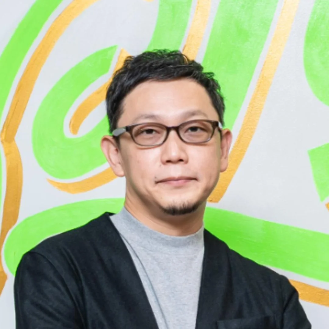 眼鏡をかけた男性が、明るい緑と金色の抽象的な背景の前で、わずかに微笑んでいます