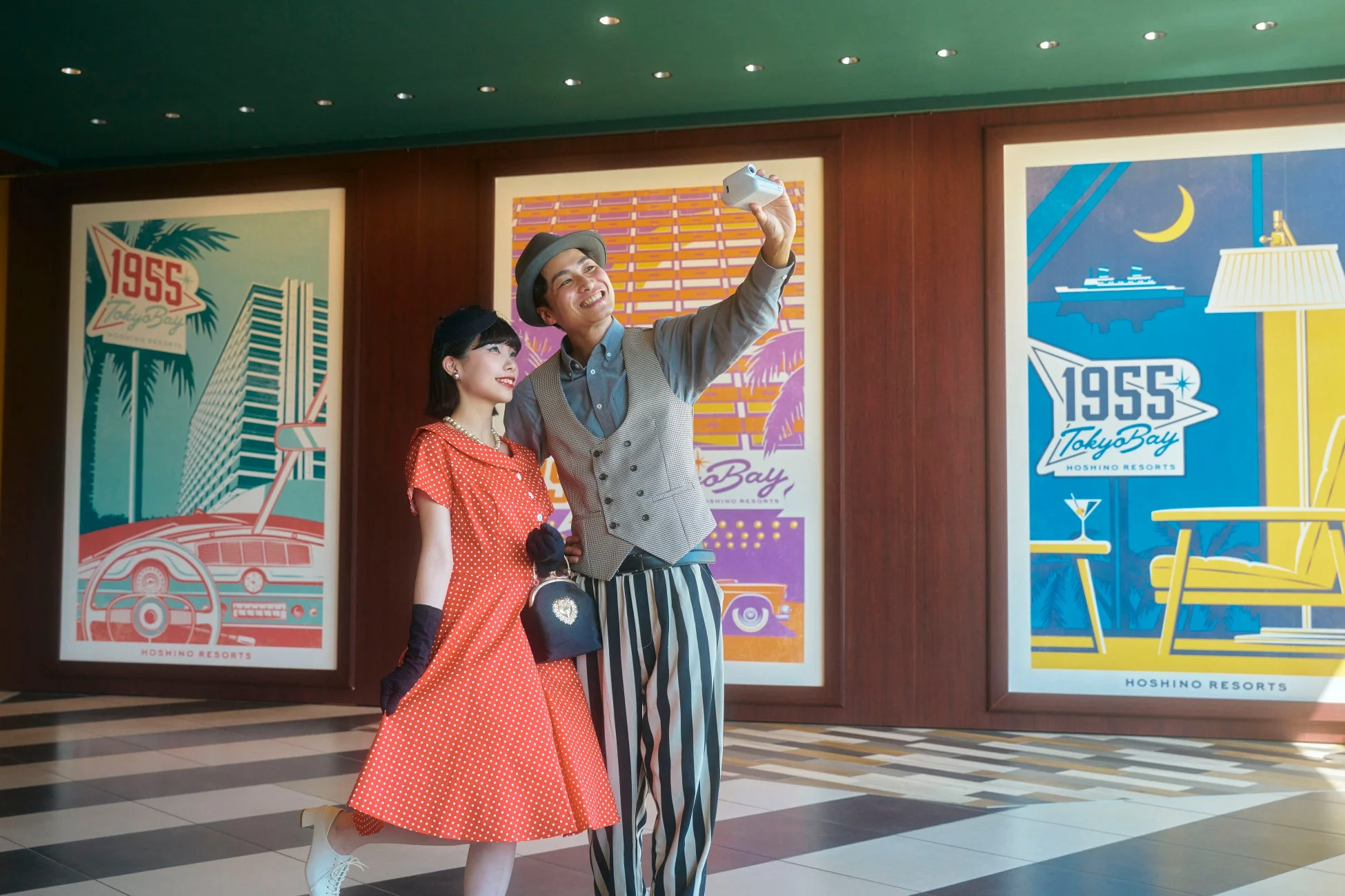 レトロな衣装を着たカップルが、ホテル「HOSHINO RESORTS 1955 Tokyo Bay」のロビーで自撮りを楽しんでいます。背景にはホテルのテーマに合わせたヴィンテージ風のポスターが飾られ、二人は笑顔で旅行の楽しい雰囲気を表現しています。