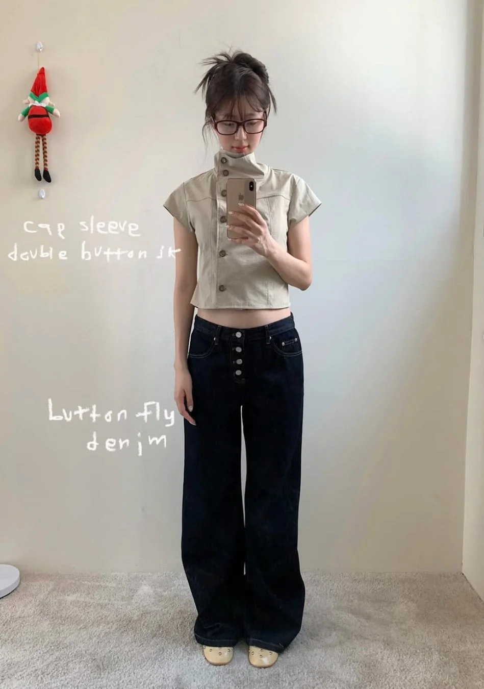 メガネをかけた女性が、ベージュのキャップスリーブダブルボタンのトップスと、ボタンフライのワイドデニムパンツを着用したコーディネートを鏡越しに撮影